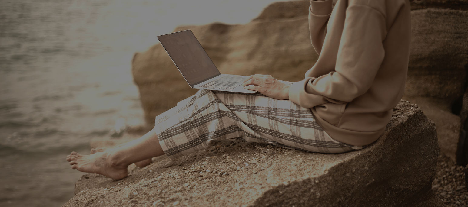 Person sitting on rocks at a beach. They are barefoot and have a laptop on their lap. Their face cannot be seen. The sea and background are blurred.