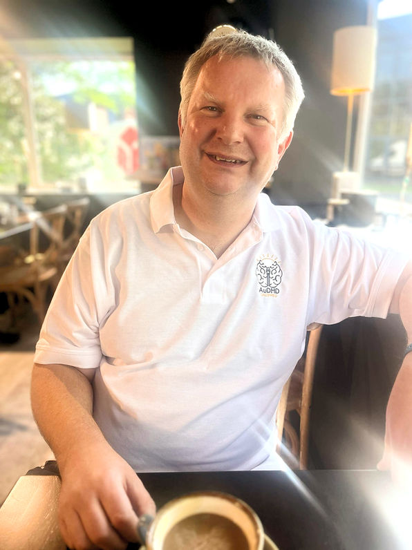 A white man sitting in a coffee table with a cup of coffee on the table. He is smiling and wearing a white polo neck t-shirt with the AuDHD Unzipped logo on it.