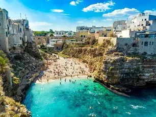 Spiaggia tra le scogliere in un centro storico della Puglia, con mare turchese, bagnanti e case in pietra sullo sfondo.