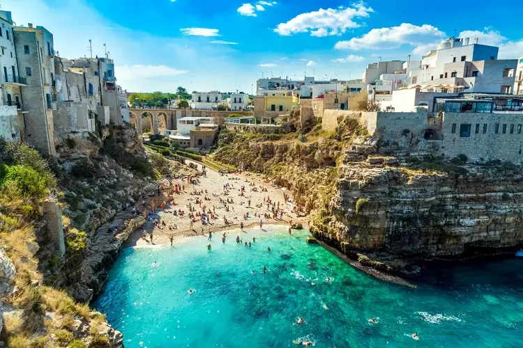 Spiaggia tra le scogliere in un centro storico della Puglia, con mare turchese, bagnanti e case in pietra sullo sfondo.