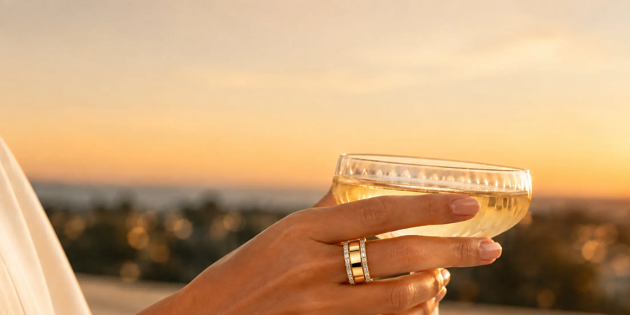 HALO Baguette in yellow gold worn over an Oura Ring, holding a coupe of champagne at sunset