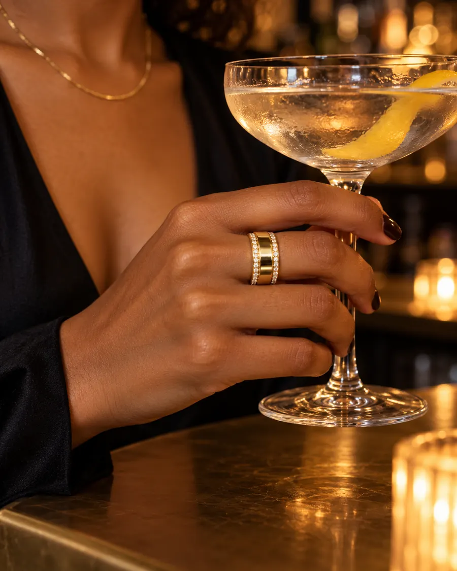 HALO Round in 14k yellow gold with round diamonds, worn over an Oura Ring while holding a coupe cocktail at a candlelit bar