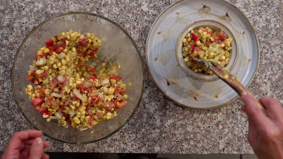 Hands serving corn salad from a glass bowl to a ceramic plate on a speckled countertop. The salad is colorful with red and yellow hues.