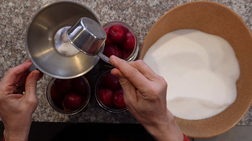Hands pour sugar into jars of red plums using a metal funnel on a granite countertop. A large bowl of sugar is beside the jars.