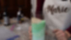Person wearing a "Marie" apron holds a glass with layered green and beige powders. Granite countertop, bottles, and bags are in the background.