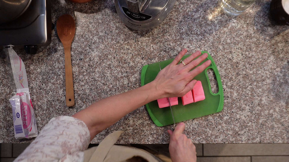 Hands slicing pink Zote bars on a green cutting board atop a speckled countertop. Nearby, a wooden spoon and food processor are visible.