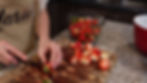 Hands slicing strawberries on a wooden board, wearing an apron with "Marie" text. Bowl of strawberries in background on a kitchen counter.