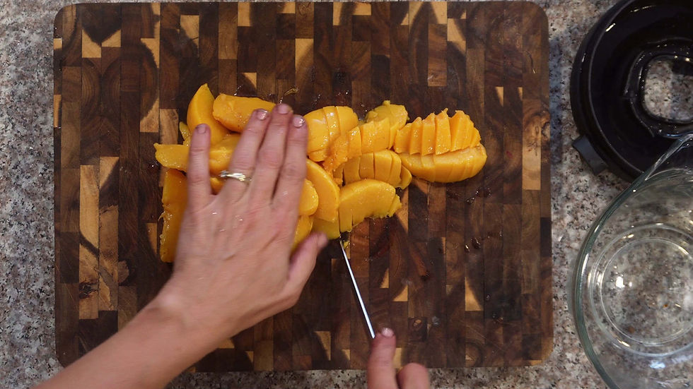 Hands slicing a ripe mango on a wooden cutting board. Kitchen countertop in the background. Clear glass bowl nearby.