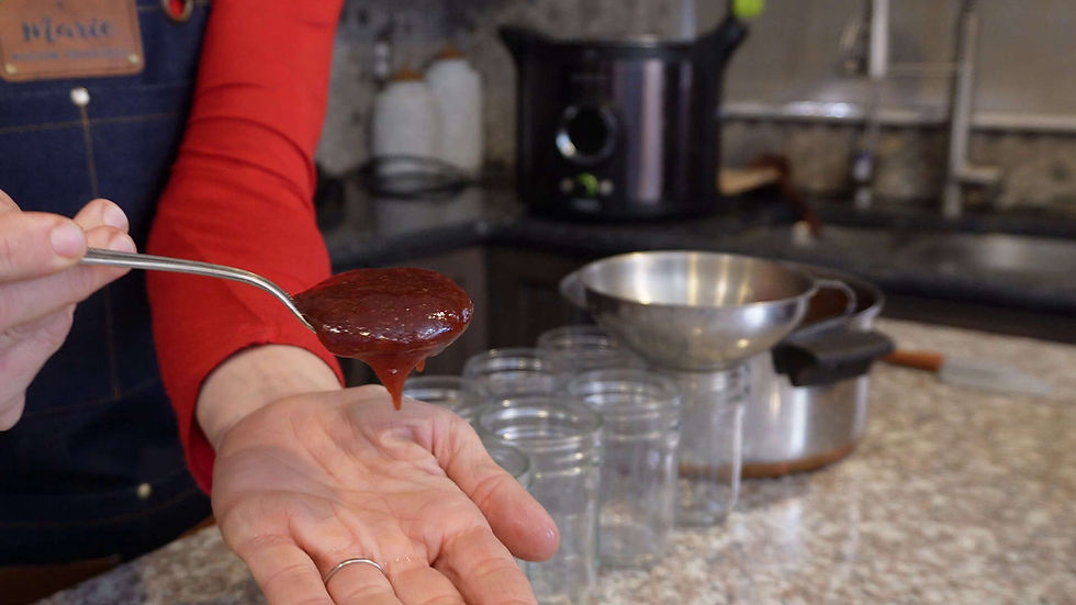 Person spooning thick red jam into a hand in a kitchen, with glass jars and a metal strainer on the counter.