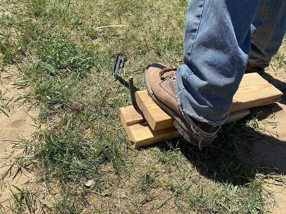 Worn work boot presses on stacked wood blocks beside a protruding rebar stake in a grassy dirt patch.