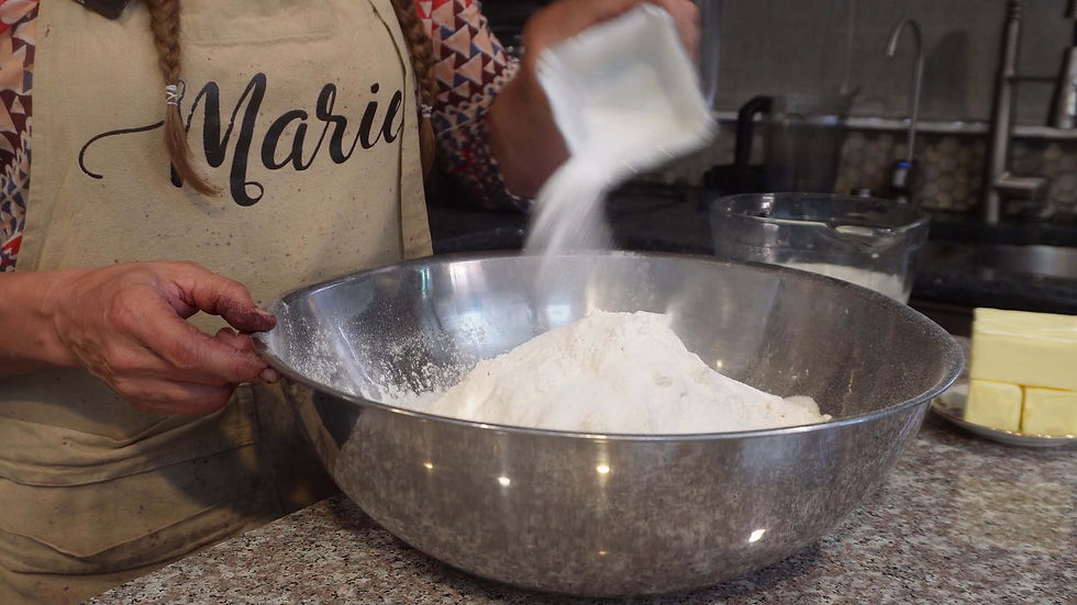 Baker pouring sugar into a large, shiny metal bowl with flour. Person wearing a patterned sleeve and apron with "Maria". Butter nearby.