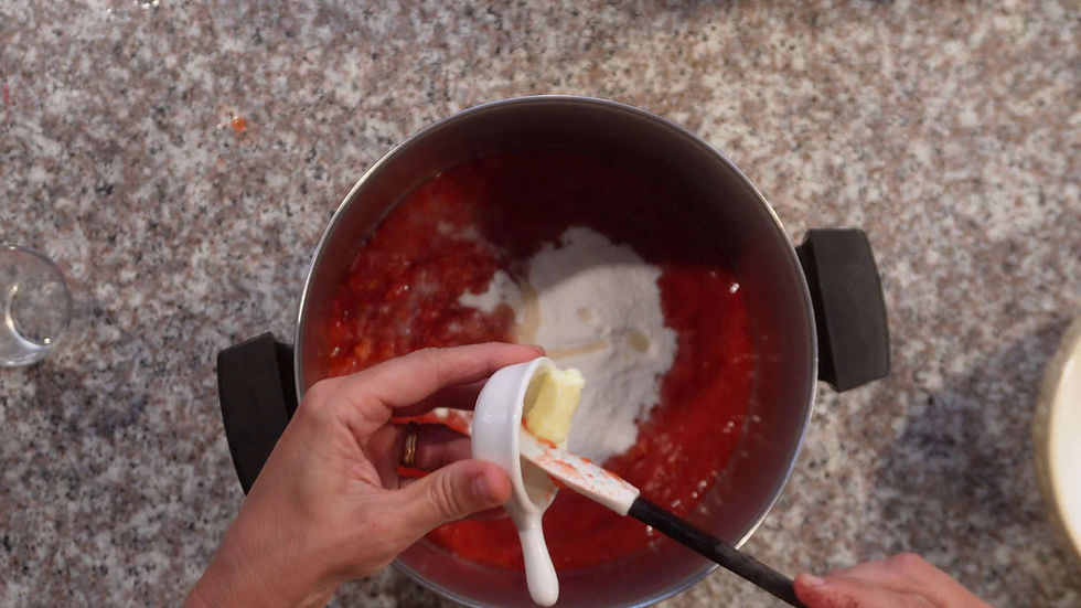 Hands adding butter to a pot with red sauce and sugar on a granite countertop. White spatula and cup visible, creating a cooking scene.