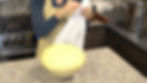 Person in apron kneads dough in a kitchen with jars, stove, and pot. Text partially reads "ar." Warm, cozy atmosphere.