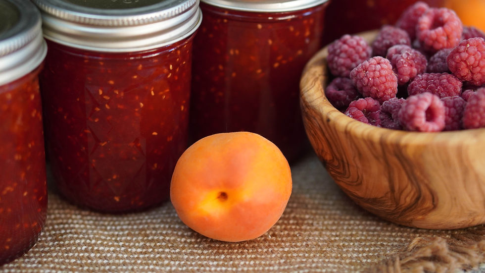 Jars of red jam, an apricot, and a wooden bowl of raspberries on a woven surface, creating a fresh and inviting scene.