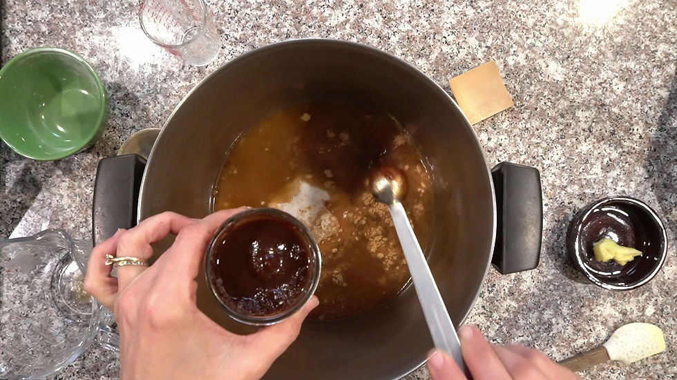 Hands mix brown sauce in a pot with spoon on a granite counter. Nearby are a green bowl, clear glass, and small dish with ginger.