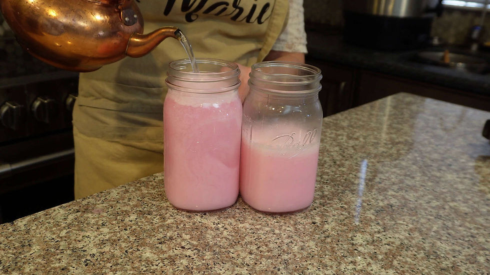 Copper kettle pouring liquid into two pink-filled jars on a granite countertop. Person in beige apron partially visible. Kitchen setting.