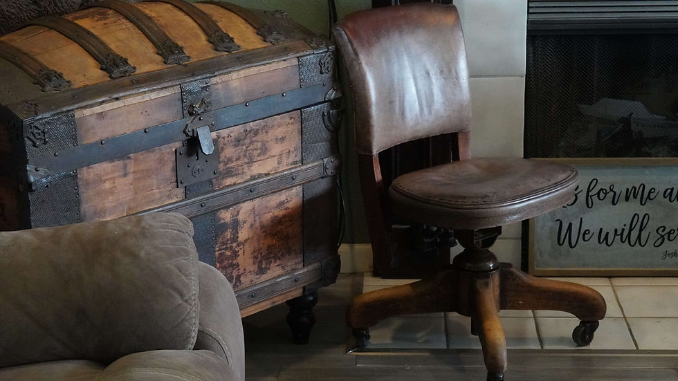 Antique wooden trunk and brown leather chair beside a fireplace. Partial view of a cozy armchair and framed text visible in the background.