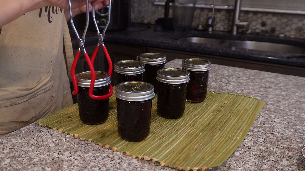 A hand uses tongs to lift a jar of dark preserves from a green mat on a granite countertop in a kitchen, with more jars in the background.