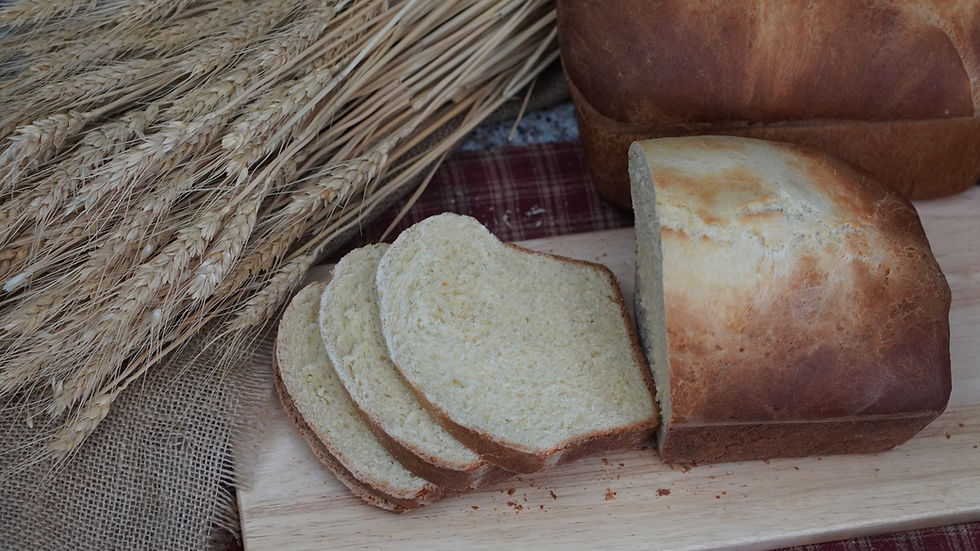 Sliced bread loaf on a wooden board with wheat stalks beside it. The setting has a rustic vibe with a burlap and plaid backdrop.
