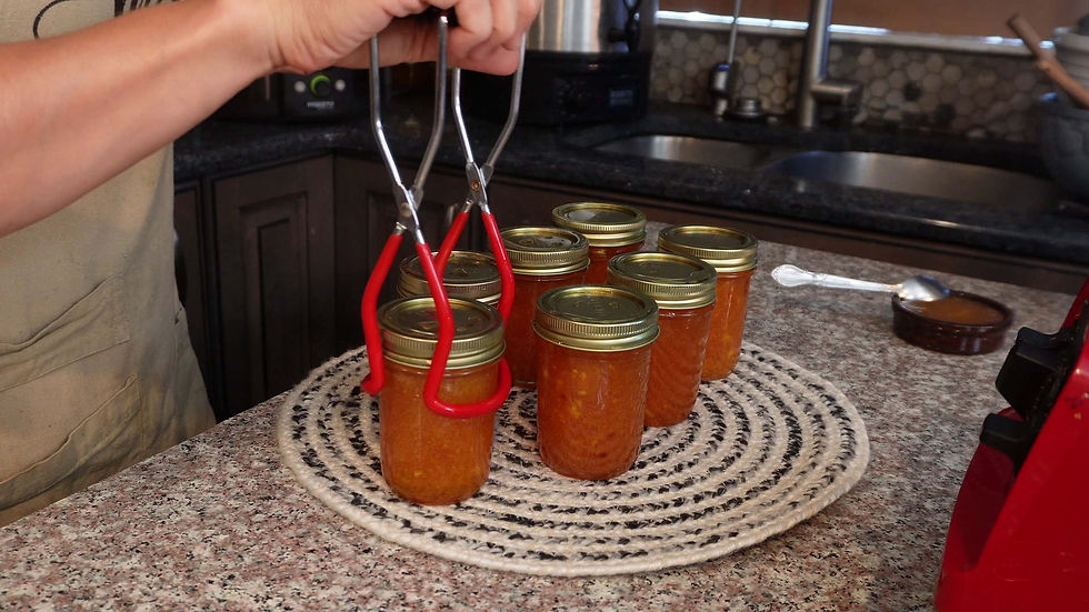 A hand uses tongs to hold a jar of nectarine jam on a woven mat, surrounded by more jars in a kitchen. The mood is orderly and homely.