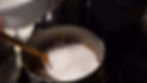 A pot with chopped apples and sugar on a stove, wooden spoon inside. A person holding a white bowl. Kitchen setting, dim lighting.