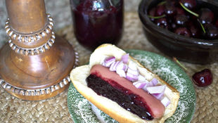 Hot dog with purple onion and dark sauce on a green plate. Wooden decor, jar, and bowl of cherries. Cozy, rustic setting.