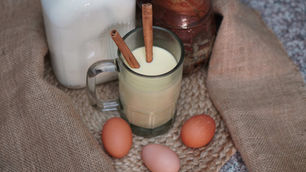 Eggnog in a glass with cinnamon sticks, surrounded by eggs, a milk bottle, and burlap fabric, creating a cozy, rustic vibe.
