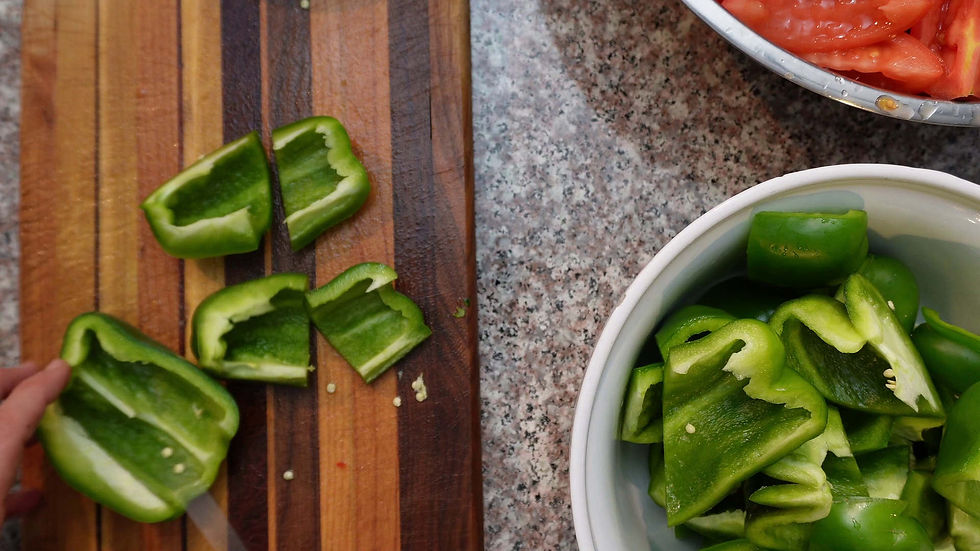 Chopped green bell peppers on a wooden board, hand reaching, with more pieces in a white bowl. Sliced tomatoes in another bowl nearby.