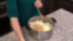 Person in a green sweater stirs chopped onions, garlic, and butter in a stainless pot on a granite kitchen counter. Cozy cooking scene.