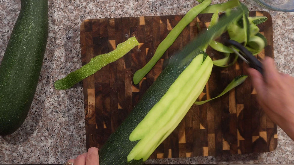 Hands peel a large zucchini on a wooden cutting board, with green peelings on a speckled kitchen counter.