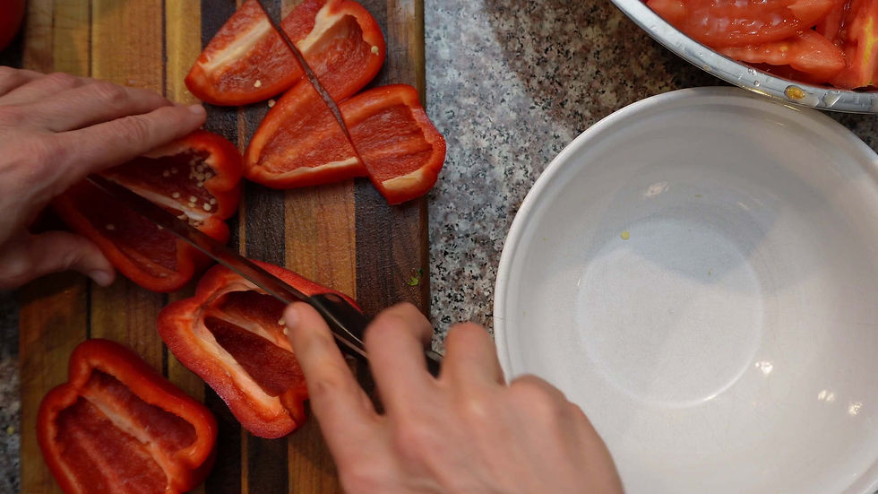 Hands slice red bell peppers on a striped cutting board. A white bowl and sliced tomatoes are nearby on a speckled countertop.