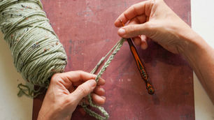 Hands crocheting pale green yarn with an orange hook on a worn pink board, a calm close-up of crafting in progress