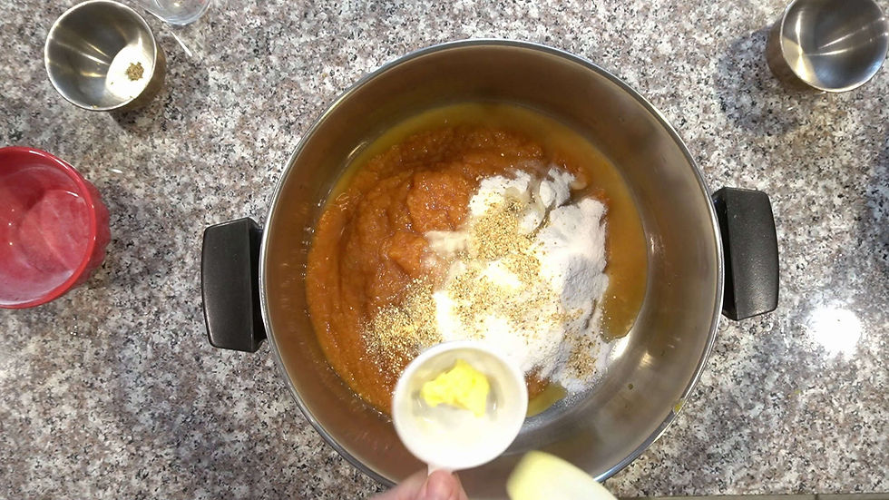 Bowl with pumpkin puree, flour, and spices on a granite countertop. A hand holds a measuring cup with butter. Steel cups and a red bowl nearby.