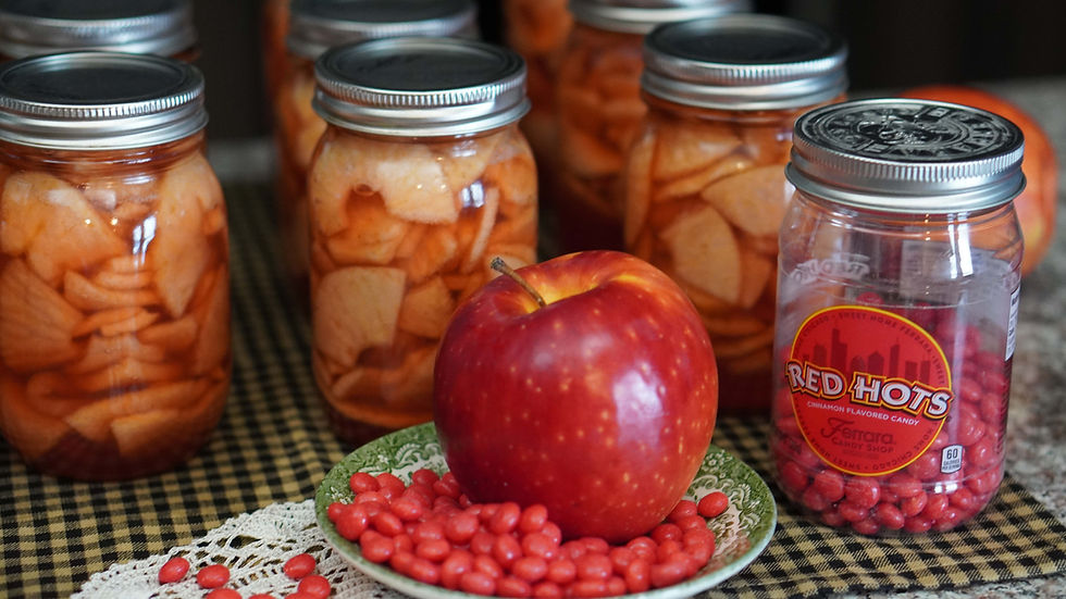 Jars filled with sliced apples in syrup, a red apple, and Red Hots candies on a checkered cloth. The mood is cozy and warm.