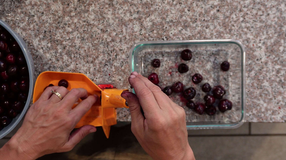 Hands using an orange cherry pitter on a speckled countertop. Cherries in a glass dish and a bowl. Hands stained with cherry juice.