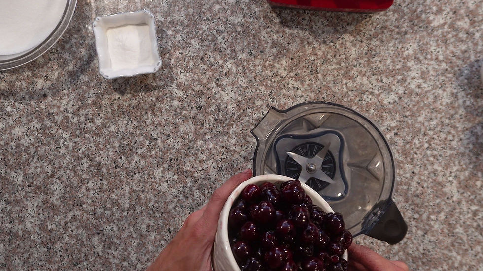 Hands holding a bowl of cherries above a blender on a speckled countertop. Nearby are dishes with sugar and baking powder.