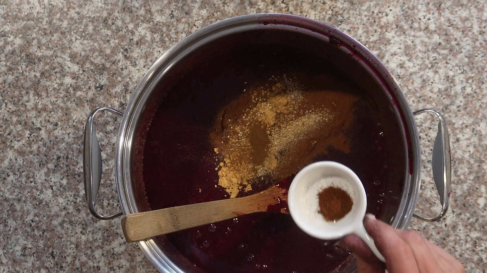 A hand holds a white cup adding spices to a pot of dark liquid on a speckled countertop. A wooden spoon rests inside the pot.