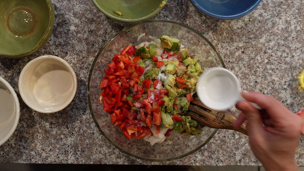 Hand sprinkles liquid into bowl with chopped red peppers, avocado, and onion on speckled countertop. Green and blue bowls surround.