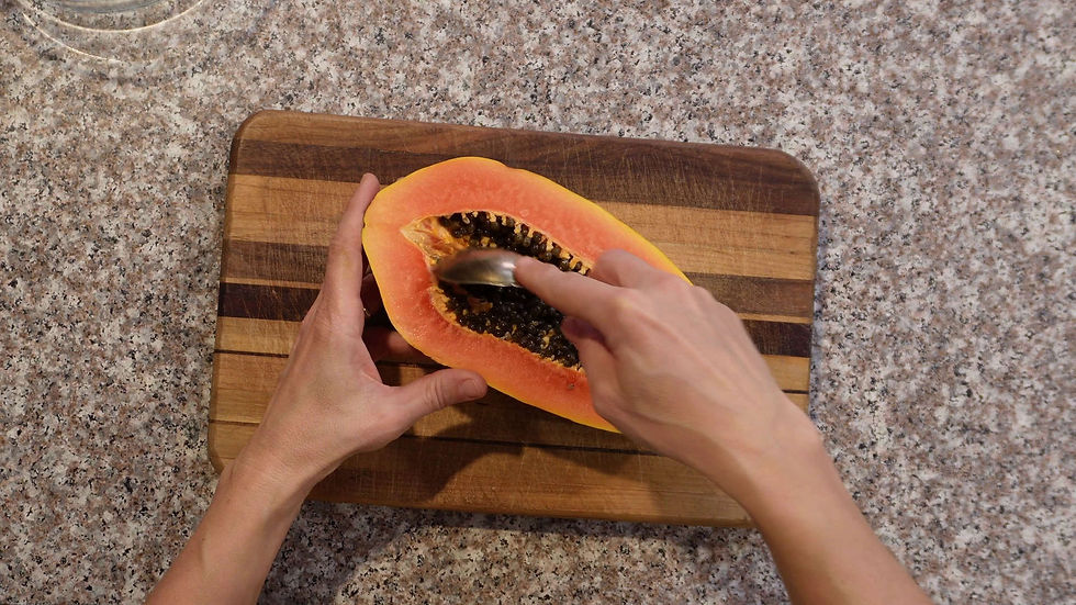 Hands scooping seeds from a halved papaya with a spoon on a wooden board. Granite countertop visible, setting is a kitchen.