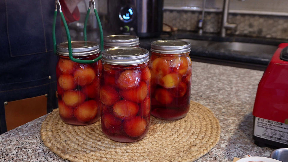Four jars of preserved red and orange fruits sit on a woven mat in a kitchen. Tongs hold one jar. A red appliance is visible on the counter.