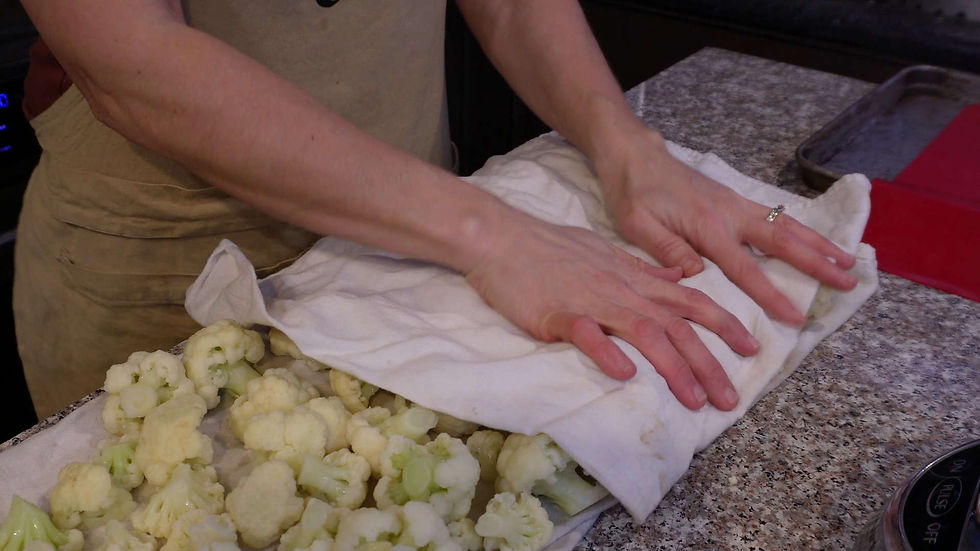 Hands pressing a white towel over cauliflower on a speckled countertop, with a baking tray nearby. The setting is a kitchen.