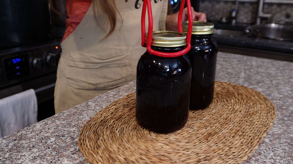 Two dark jars with golden lids on a woven mat in a kitchen. A person in an apron holds a red jar lifter. Oven shows "5:54".