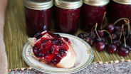 Plate with toast topped with cherry jam beside fresh cherries and jars of jam, on a green mat. Rustic and appetizing setting.