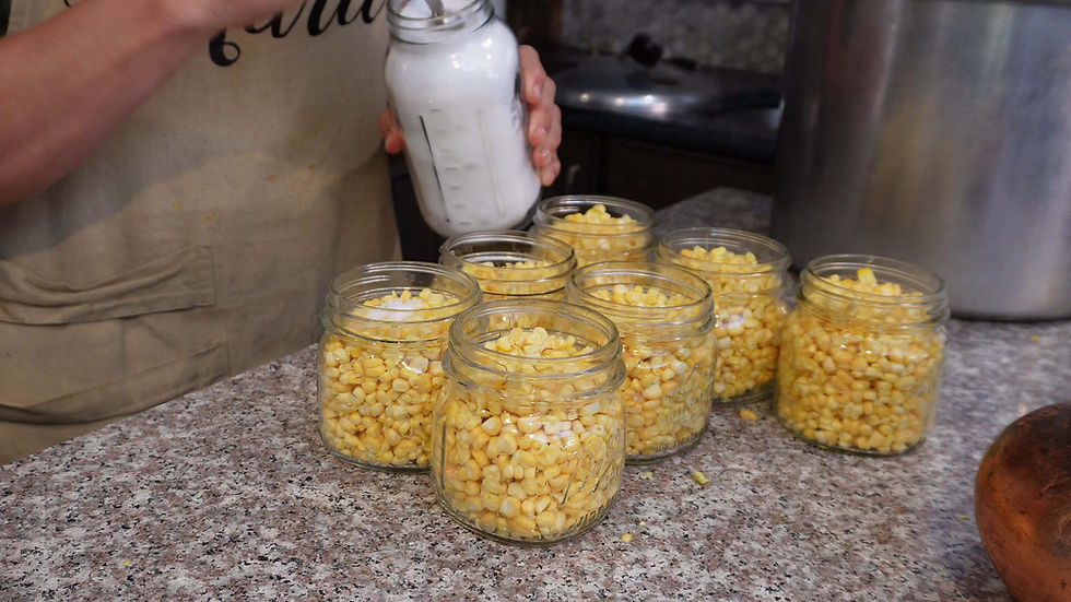 Person in apron fills jars of corn with salt on a kitchen counter. Seven jars are visible, each partially filled with yellow corn.