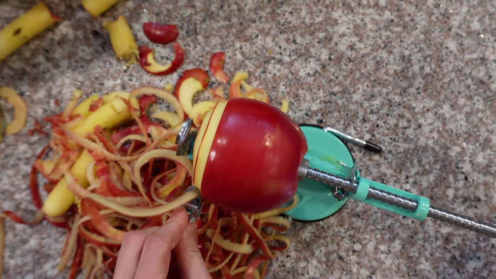 Red apple being peeled with a teal apple peeler on a speckled countertop. Apple peels and cores are scattered around, hand adjusting the peeler.