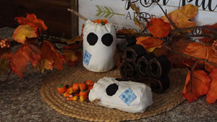 Fabric ghost decorations and candy corn on a woven mat with autumn leaves. "Boo" sign and leafy background create a festive, cozy vibe.