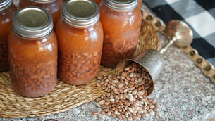 Jars of beans sit on a woven mat next to a metal cup spilling beans onto a speckled countertop. Black and white cloth partially visible.