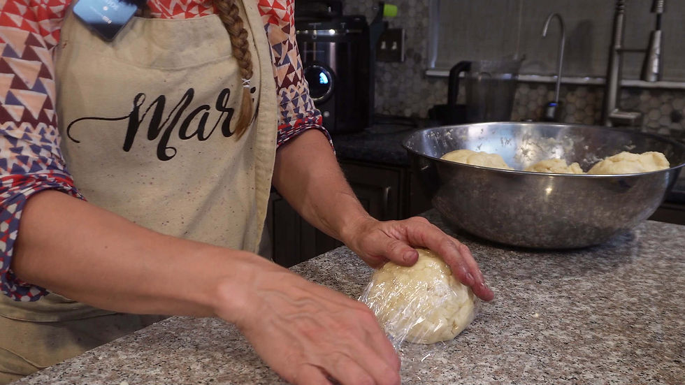 Person in patterned apron wraps dough in plastic on marble counter, with bowl of dough in kitchen background. Apron has "Mari" text.