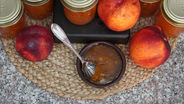 Jars of nectarine jam, fresh nectarines, and a small bowl with a spoon on a woven mat on a granite countertop, creating a homely feel.
