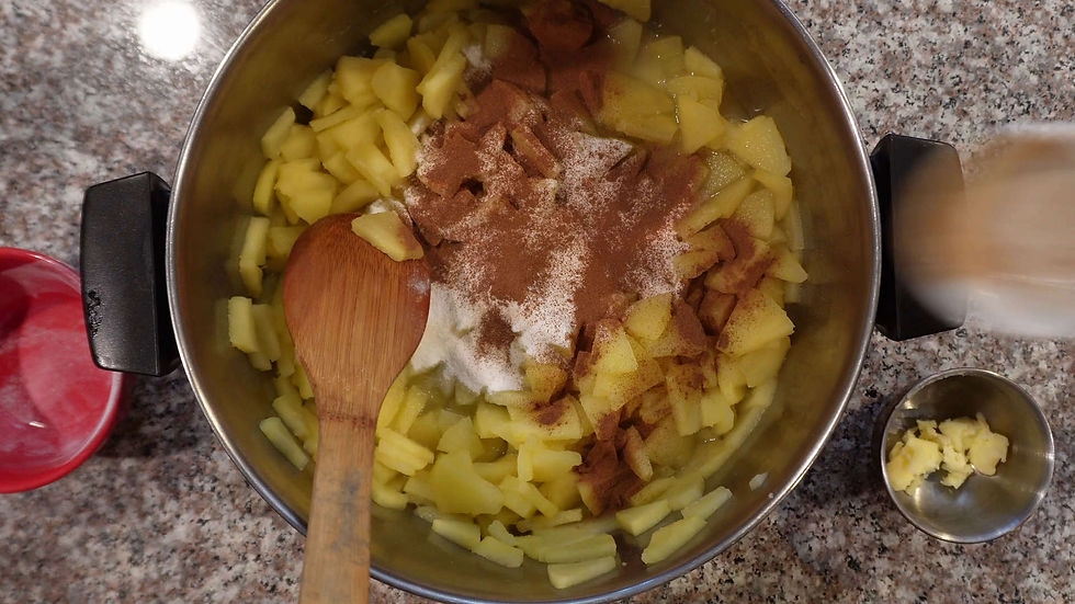 Chopped apples, cinnamon, and sugar in a pot with a wooden spoon on a granite countertop; red bowl and butter in small bowl nearby.